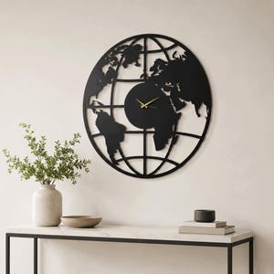 Puede incluir: Un gran reloj de pared circular con un marco de metal negro en forma de mapa mundial con líneas de longitud y latitud. La esfera del reloj es de color negro sólido con manecillas finas de color dorado para las horas y los minutos.