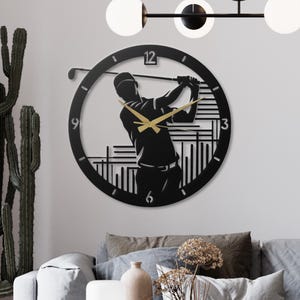 May include: Black metal golf-themed wall clock. The clock features a silhouette of a golfer mid-swing, with gold-coloured clock hands. The clock face has the numbers 3, 6, 9, and 12. The clock is mounted on a white wall.