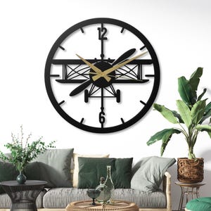 May include: A black metal wall clock featuring a biplane design within a circular frame. The clock has gold-colored hands and a propeller. The numbers 12 and 6 are clearly marked. The clock is mounted on a white wall, above a sofa.