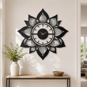 May include: A large, circular wall clock featuring a black, stylised lotus flower design with pointed petals. The centre has a solid black circular face with thin gold hands, surrounded by a white ring with black hour markers and a horizontal bar.