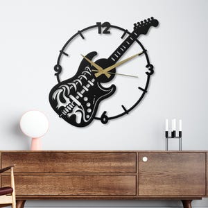 May include: A black guitar-shaped wall clock with gold hands and a circular clock face. The guitar design features a skull motif on the body. The clock is mounted on a white wall above a wooden cabinet.