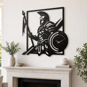 May include: A black metal wall clock featuring a silhouette of a Spartan warrior in armor holding a spear. The warrior is framed within a square border, and a circular clock face with thin gold hands is integrated into the design on the right side.