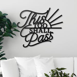 Op de afbeelding: Zwarte metalen wandkunst met de tekst "This Too Shall Pass" in een decoratief script. Het ontwerp bevat een gestileerd zonmotief en is bevestigd aan een witte muur boven een witte bank.