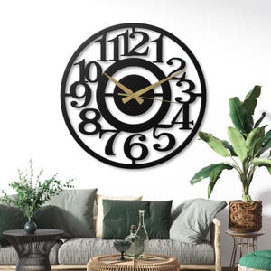 May include: A large, black, circular wall clock with cut-out numbers and a gold-colored clock hands. The clock is mounted on a white wall. The clock's diameter is approximately 24 inches.
