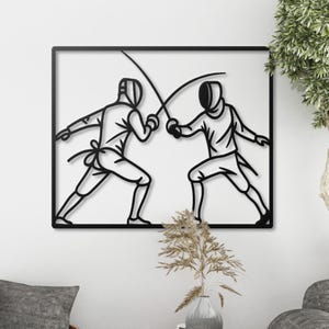 May include: Black metal wall art showing two fencers duelling, set within a rectangular frame. The silhouettes depict fencers in protective gear, holding swords, engaged in a fencing match. The artwork is mounted on a white wall.