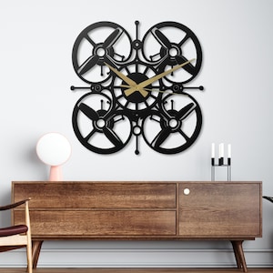 May include: A black metal wall clock with a drone propeller design. The clock has four propeller-shaped elements around a central clock face with gold hands. It's mounted on a white wall above a wooden cabinet. The clock's dimensions are approximately 40 cm x 40 cm.