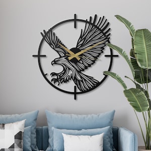 May include: A black metal wall clock featuring an eagle design. The clock has a circular frame with a stylized eagle in the center, and gold-colored clock hands. The clock is mounted on a light gray wall, with a blue sofa and a plant in the background.