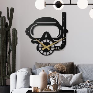 Op de afbeelding: Een zwarte metalen wandklok in de vorm van een duikmasker en snorkel. De wijzers zijn goudkleurig en de wijzerplaat heeft een gestileerd ontwerp. De klok is bevestigd aan een witte muur boven een grijze bank.