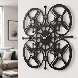 Könnte beinhalten: Eine schwarze Wanduhr mit einem zentralen runden Zifferblatt und goldfarbenen Zeigern. Der Rahmen hat ein symmetrisches Design mit vier großen kreisförmigen Aussparungen, die jeweils eine propellerartige Struktur wie bei einer Drohne enthalten.