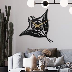 May include: A black metal wall clock shaped like a manta ray. The clock face is circular with Roman numerals and gold hands. The manta ray design has cut-out details and is mounted on a white wall, adding a unique decorative element to the room.