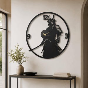 Peut inclure: Une horloge murale circulaire noire représentant la silhouette d'un samouraï tenant un sabre. Le cadran comporte les chiffres 12, 3 et 9 dans une police grasse, avec des aiguilles dorées fines pour les heures et les minutes au centre.