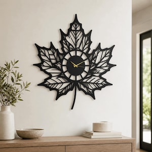 May include: A black wall clock shaped like a maple leaf with intricate cut-out vein details. The centre features a circular clock face with thin gold-coloured hour and minute hands. The design is symmetrical with a pointed stem at the bottom.