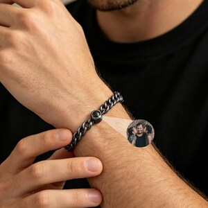 Op de afbeelding: Een donkergrijze kettingarmband met een cirkelvormige sluiting. De armband wordt om de pols van een persoon gedragen. Een kleine inset afbeelding toont een groep mensen.
