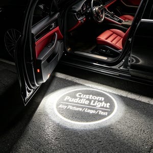 May include: A car door open to reveal a bright circular projection on the ground displaying the text Custom Puddle Light and Any Picture / Logo / Text. The vehicle interior features black and red leather seating and a steering wheel.