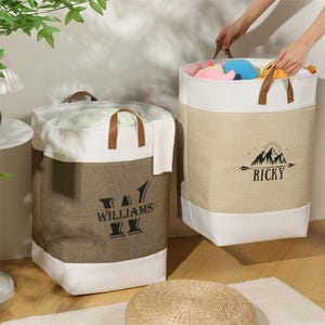 May include: Two canvas laundry hampers with leather handles. One is brown with "WILLIAMS" printed on it, the other is tan with "RICKY" and a mountain graphic. Both have white tops and bottoms.