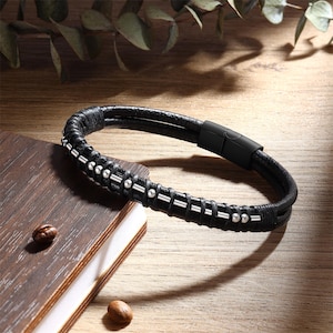 Puede incluir: Pulsera de cuero negro con cuentas y detalles plateados. La pulsera tiene un cierre magnético y se muestra sobre una superficie de madera. El diseño incorpora múltiples hebras de cuero para una apariencia en capas.