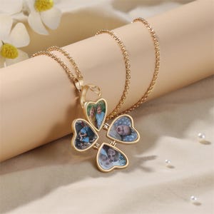 May include: A gold-coloured pendant necklace featuring a four-leaf clover design. Each leaf opens to display a small photograph. The necklace hangs on a gold chain, presented on a beige surface with white flowers.