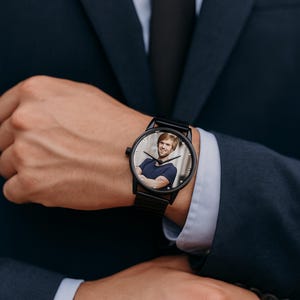 May include: Black wristwatch with a round face displaying a colour photograph of a person. The watch has a black strap and case. The person in the photograph is wearing a blue top. The watch is on a person's wrist.