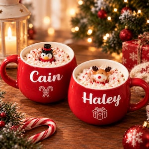 Könnte beinhalten: Zwei rote Keramiktassen gefüllt mit Schlagsahne und Pfefferminzstücken. Die linke Tasse zeigt den Namen Cain und ein Zuckerstangen-Symbol. Die rechte Tasse zeigt den Namen Haley und ein Geschenk-Symbol. Eine kleine Pinguinfigur sitzt in der linken Tasse und eine Rentierfigur in der rechten.