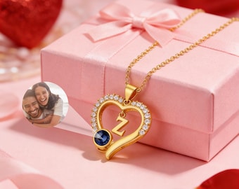 May include: A gold-coloured heart-shaped pendant necklace with the letter "Z" and a blue gemstone. The pendant is encrusted with small, clear crystals and hangs on a delicate chain. A pink gift box with a bow and red heart decorations are in the background.