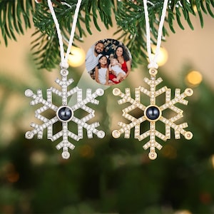 Peut inclure: Deux pendentifs en forme de flocon de neige, l'un argenté et l'autre doré, avec une photo de famille au centre. Le pendentif argenté est orné de cristaux transparents, le doré de cristaux transparents. Les deux pendentifs sont suspendus à un cordon blanc.
