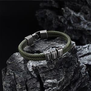 Può includere: Un braccialetto verde intrecciato con tre perline d'argento con i nomi "Loana", "Martin" e "Fabian". Il braccialetto ha una chiusura argentata ed è esposto su una superficie rocciosa strutturata grigio scuro.