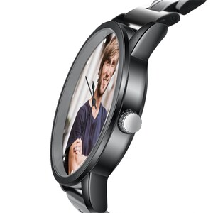 May include: A black wristwatch with a stainless steel strap. The watch face displays a colour photograph of a person. The watch has a silver crown and black hands. The watch is angled to show the face and the strap.
