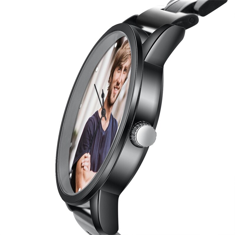 May include: A black wristwatch with a stainless steel strap. The watch face displays a colour photograph of a person. The watch has a silver crown and black hands. The watch is angled to show the face and the strap.