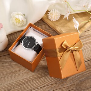 May include: A black-faced wristwatch with a silver bezel and black leather strap sits in an open orange gift box with a white cushion. A matching closed gift box with a gold ribbon is next to it. The watch is a stylish accessory.