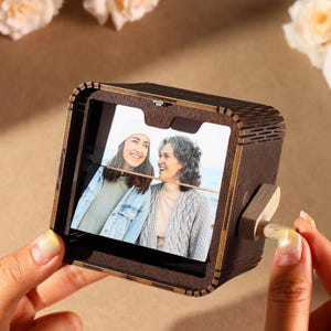 Peut inclure: Un cube photo en bois marron foncé avec un mécanisme rotatif, affichant une photographie de deux femmes souriantes. Le cube a une petite poignée en bois pour tourner. La photo est visible à travers une ouverture rectangulaire.