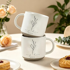 May include: Two white ceramic mugs, each with a grey base, a curved handle, and a black botanical design, stacked on top of each other. The mugs feature the initials "j + h" and the date "02.15.25".