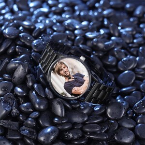 May include: A black wristwatch with a stainless steel strap and a round face. The watch face displays a colour photograph of a person. The watch is resting on a bed of dark, smooth stones.