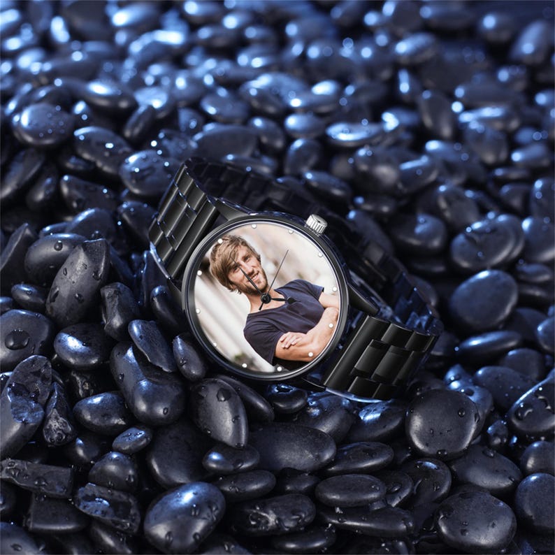 May include: A black wristwatch with a stainless steel strap and a round face. The watch face displays a colour photograph of a person. The watch is resting on a bed of dark, smooth stones.