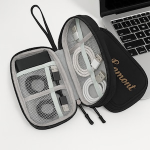 May include: Black zippered travel case with multiple compartments, holding a black power bank, black USB cables, and white USB cables. The case is open, revealing the contents. A laptop is in the background.