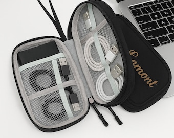 Può includere: Custodia da viaggio nera con cerniera e più scomparti, contenente un power bank nero, cavi USB neri e cavi USB bianchi. La custodia è aperta, rivelando il contenuto. Un laptop è sullo sfondo.