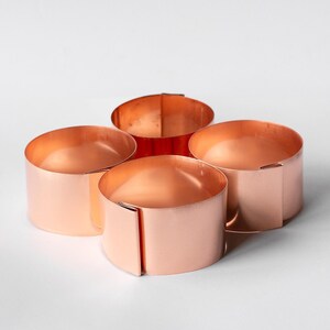May include: Four copper-coloured metal napkin rings on a white surface. Each ring is cylindrical with a folded seam. The rings have a shiny, reflective surface, and are designed for table settings.