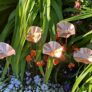 May include: Five copper garden ornaments in the shape of stylized flowers. The metallic flowers are set among green foliage, orange and red flowers, and small blue flowers. The copper flowers have a reflective, shiny surface.