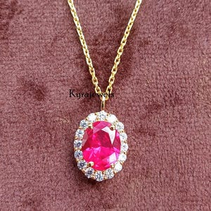 Könnte beinhalten: Eine goldfarbene Halskette mit einem Anhänger, der einen großen, ovalförmigen, leuchtend roten Edelstein zeigt, der von kleineren weißen und blauen Edelsteinen umgeben ist. Die Kette liegt auf einem weinroten Hintergrund.