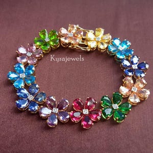 May include: A gold bracelet featuring flower-shaped gemstones in various colors, including blue, green, pink, and orange. Each flower has a small diamond in the center. The bracelet is on a brown background, and the text "Kyrajewels" is visible.