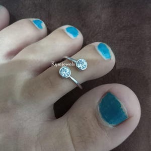 May include: A silver toe ring with two clear, round gemstones. The ring is designed to wrap around two toes. The image shows the ring on a foot with blue nail polish. The text "Kyrajewels" is visible.