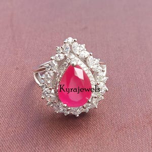 May include: A silver ring featuring a large, pear-shaped, bright pink gemstone surrounded by smaller, clear stones. The ring has a detailed, ornate setting. The text "Kyrajewels" is visible on the ring.