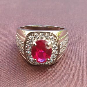 Könnte beinhalten: Ein silberner Ring mit einem großen, oval geschliffenen Rubin in der Mitte, umgeben von kleineren, klaren Edelsteinen. Der Ring hat ein klassisches, elegantes Design.