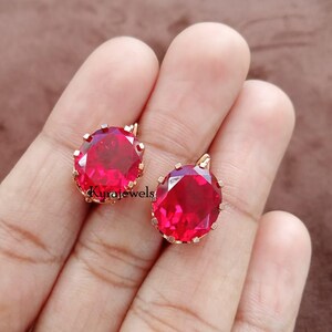 May include: Two gold-toned pendant earrings, each featuring a large, oval-cut, ruby-red gemstone. The gemstones are set in a gold-colored metal with a decorative, pronged setting. The earrings are displayed on a brown background.