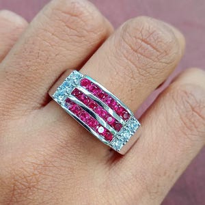 May include: A silver ring with a rectangular face, set with three rows of red gemstones and two rows of white gemstones. The ring is on a person's finger, with a blurred background.