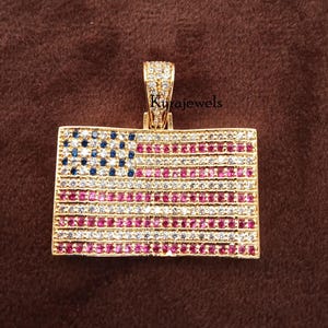May include: Gold-coloured pendant in the shape of the American flag, with blue, red, and white gemstones. The pendant is rectangular with a bail at the top. The text "Kurajewels" is visible on the pendant.
