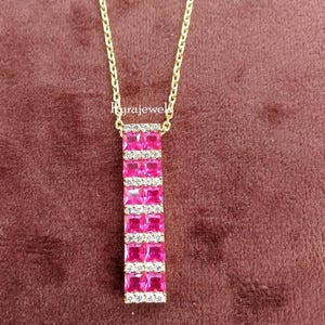 May include: A gold-colored necklace with a rectangular pendant. The pendant features rows of bright pink square-cut gemstones, separated by rows of smaller, clear gemstones. The necklace hangs on a gold chain against a brown background.