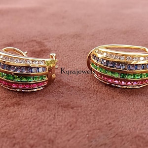 Puede incluir: Un par de pendientes de aro dorados. Cada pendiente presenta filas de piedras preciosas rosas, verdes, azules y blancas. Los pendientes tienen un diseño curvo y un cierre seguro.