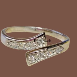 Pode incluir: Um anel prateado com um design dividido. Cada lado do anel é adornado com pequenas pedras preciosas transparentes. O anel tem o texto "Kyrajewels" na lateral.