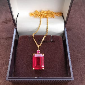 May include: A gold-colored necklace with a rectangular, red gemstone pendant. The pendant is set in a gold frame and has a small, round red gemstone above it. The necklace is displayed in a brown jewelry box. The text "Karajewels" is visible.