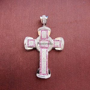 May include: A silver cross pendant, adorned with pink and clear gemstones. The cross has an intricate design, a loop for a chain, and the text "Kyrajewels". The pendant is displayed against a maroon backdrop.